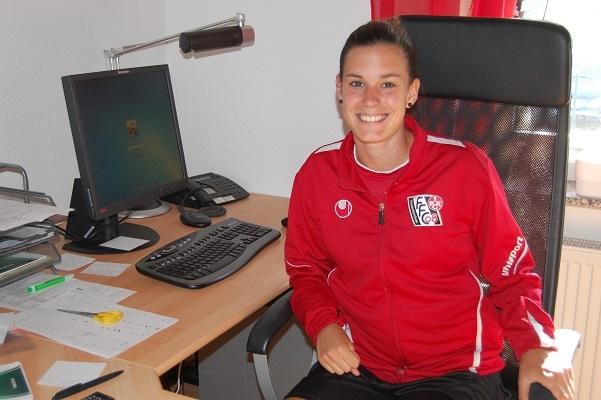 L&auml;chelnde Person in roter Sportjacke sitzt an einem Schreibtisch mit Computer und B&uuml;romaterialien.