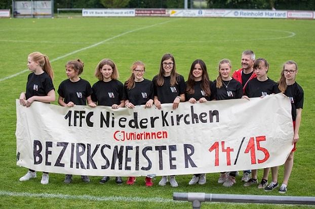 Gruppe junger Frauen in schwarzen T-Shirts h&auml;lt ein transparents Banner mit der Aufschrift &bdquo;Bezirkmeister 14/15&ldquo; auf einem Fu&szlig;ballfeld.