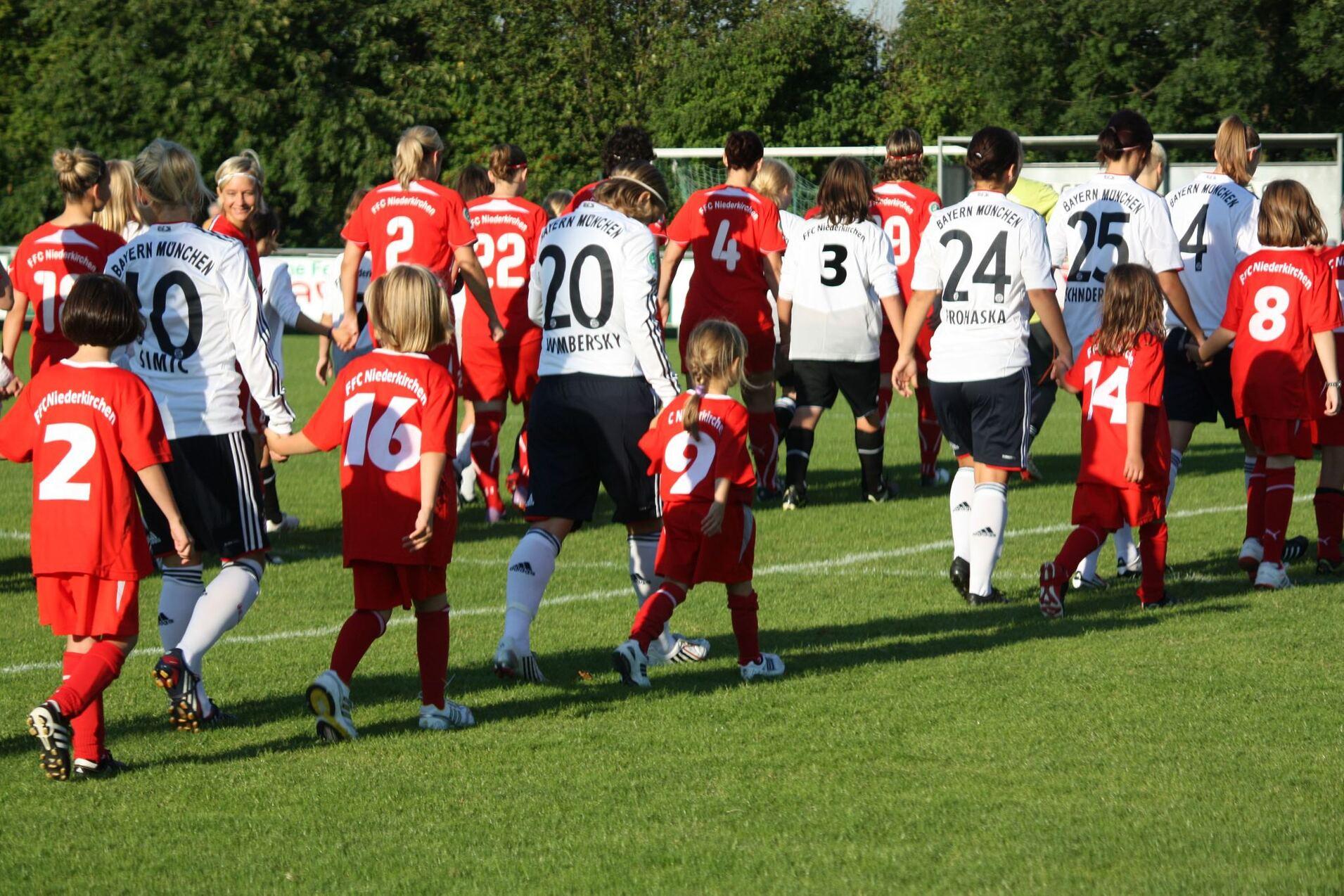 Spielerinnen beider Fußballmannschaften sowie Kinder in Trikots laufen gemeinsam auf dem Spielfeld zur Zuschauertribüne.