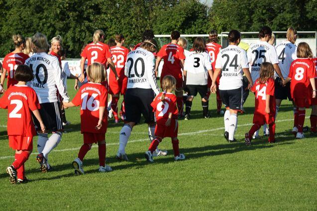 Spielerinnen beider Fu&szlig;ballmannschaften sowie Kinder in Trikots laufen gemeinsam auf dem Spielfeld zur Zuschauertrib&uuml;ne.