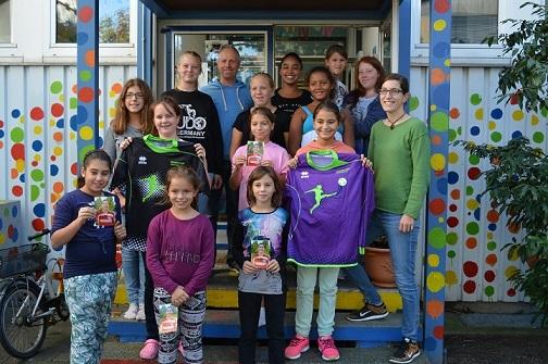 Gruppe von M&auml;dchen und Betreuern steht vor einem farbenfrohen Eingang, h&auml;lt Preise und sportliche Jerseys in den H&auml;nden.