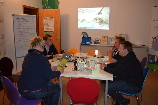 Vier Personen sitzen an einem Tisch, w&auml;hrend Pr&auml;sentationsmaterial an der Wand gezeigt wird.