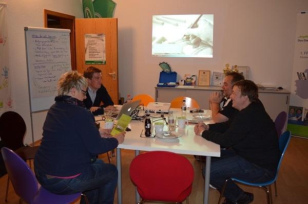 Vier Personen sitzen an einem Tisch, w&auml;hrend Pr&auml;sentationsmaterial an der Wand gezeigt wird.