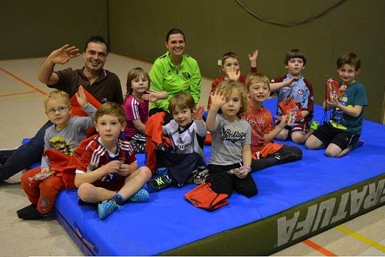 Gruppe von Kindern und zwei Erwachsenen auf einer blauen Matte, alle l&auml;cheln und winken in Richtung Kamera.