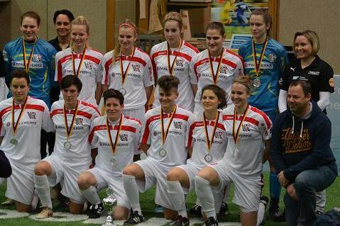 Mannschaft aus sechs Frauen und fünf Männern in weißen Trikots mit Medaillen, posiert vor einem Sporthintergrund.