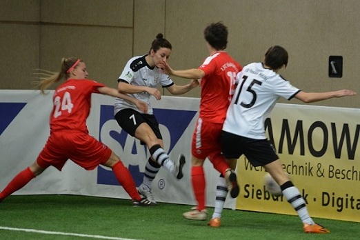 Zwei Spielerinnen in roten Trikots dr&uuml;cken eine Spielerin im wei&szlig;en Trikot gegen die Bande auf einem Fu&szlig;ballfeld.