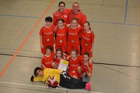 Mädchenfußballmannschaft in roten Trikots posiert stolz mit einer Urkunde in einer Sporthalle.