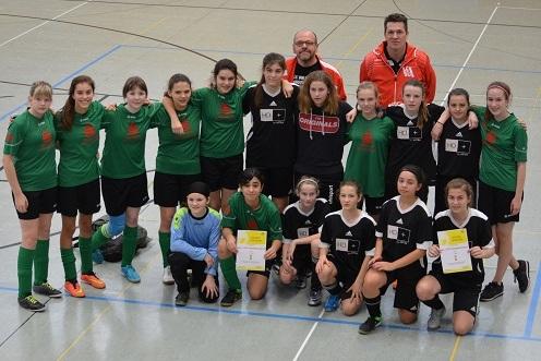 Gruppenfoto von zwei Mädchenfußballteams mit Pokalen in einer Sporthalle, Trainer im Hintergrund. Alle lächeln in die Kamera.