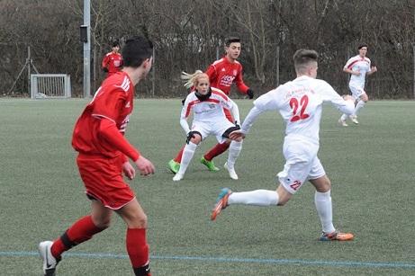 Zwei Fu&szlig;ballspieler in roten und wei&szlig;en Trikots spielen auf einem Kunstrasenplatz; eine Spielerin ist im Hintergrund.