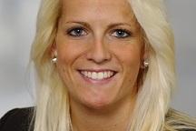 Porträt einer lächelnden Frau mit blonden Haaren, die einen schwarzen Blazer trägt. Heller Hintergrund.