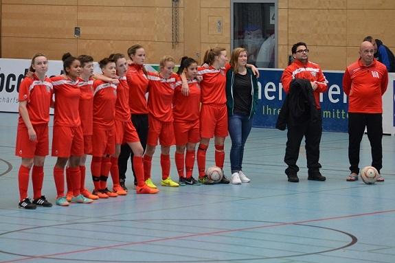 Mannschaft in roten Trikots steht auf einem Hallenfu&szlig;ballfeld, umgeben von Trainern, mit einem Ball auf dem Boden.