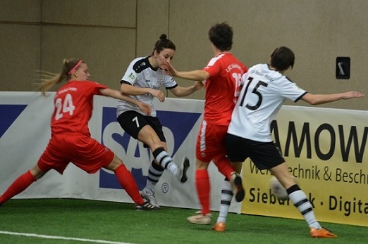 Zwei Spielerinnen in roten Trikots dr&uuml;cken eine Spielerin im wei&szlig;en Trikot gegen die Bande auf einem Fu&szlig;ballfeld.