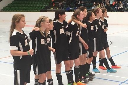 Eine M&auml;dchenfu&szlig;ballmannschaft in schwarzen Trikots steht aufgereiht in einer Sporthalle und blickt nach vorne.