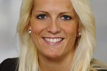 Portr&auml;t einer l&auml;chelnden Frau mit blonden Haaren, die einen schwarzen Blazer tr&auml;gt. Heller Hintergrund.