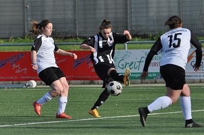 Zwei Spielerinnen in weißen Trikots und eine Spielerin in schwarz-weißer Kleidung kämpfen um den Ball auf dem Fußballfeld.