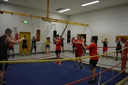 Gruppe junger Boxer in rote Trainingsshirts trainiert in einem Ring mit Boxsäcken und Seilabsperrungen.