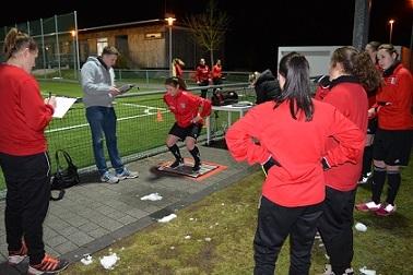 Gruppentraining im Fu&szlig;ball: Spielerin testet auf einer Plattform, w&auml;hrend Trainer und Teamkolleginnen zuschauen.