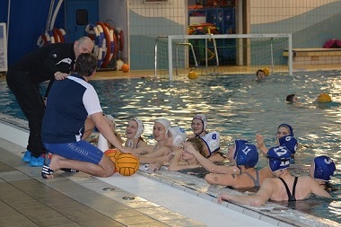 Wasserballtrainer erkl&auml;rt Technik einer Gruppe von Spielerinnen am Beckenrand w&auml;hrend des Trainings in einem Hallenbad.