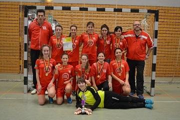 Gruppenfoto eines Handballteams in roten Trikots mit Medaillen, Pokal und Urkunde, Trainer stehen hinter den Spielerinnen.