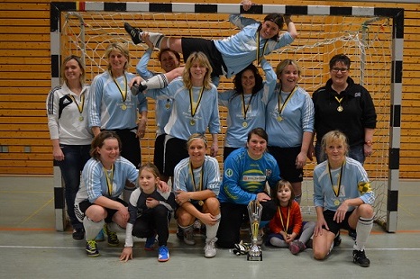 Frauenfu&szlig;ballteam mit Medaillen und Pokal posiert vor einem Tor in einer Sporthalle. Einige Mitglieder in blauen Trikots.