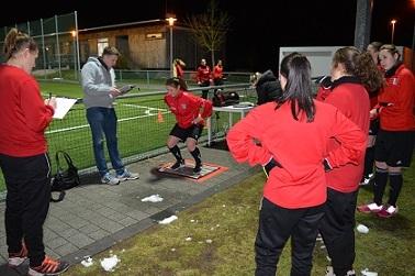 Gruppentraining im Fu&szlig;ball: Spielerin testet auf einer Plattform, w&auml;hrend Trainer und Teamkolleginnen zuschauen.