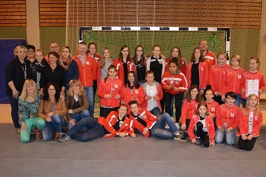 Gruppenshot von M&auml;dchen- und Damenmannschaft in roten Trainingsanz&uuml;gen, stehen und sitzen vor einem Handballtor.