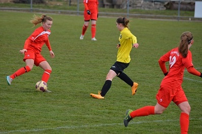 Drei Fu&szlig;ballspielerinnen auf einem Rasenfeld: eine in rotem, zwei in gelbem und schwarzem Trikot spielen den Ball.
