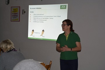 Frau pr&auml;sentiert Informationen &uuml;ber Fu&szlig;ball vor Publikum, mit einer Folie im Hintergrund.