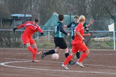 Spielszene eines Fu&szlig;ballspiels mit Spielern in roten und gr&uuml;nen Trikots auf einem braunen Rasenplatz.