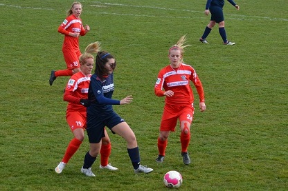 Zwei Spielerinnen in roten Trikots greifen eine Spielerin in blauem Trikot am Fu&szlig;ballfeld an. Der Ball liegt am Boden.