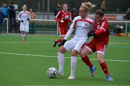 Zwei Fu&szlig;ballspielerinnen in Aktion auf einem Kunstrasenplatz, eine in wei&szlig;em und die andere in rotem Trikot.