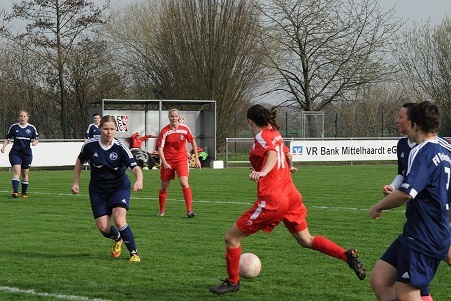 Zwei Fu&szlig;ballmannschaften spielen auf einem Rasenplatz, eine Spielerin in Rot dribbelt den Ball, w&auml;hrend andere reagieren.