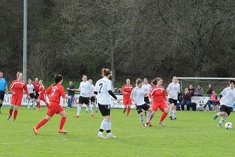 Frauenfu&szlig;ballspiel mit Spielerinnen in roten und wei&szlig;en Trikots auf einem Rasenplatz, Zuschauer im Hintergrund.