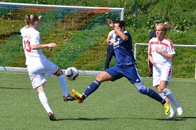 Zwei Fu&szlig;ballspielerinnen im Wettkampf um den Ball auf einem Kunstrasenplatz, eine Spielerin in Blau, die andere in Wei&szlig;.