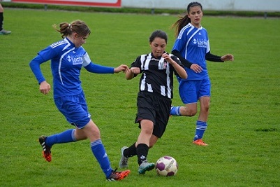 Drei Fu&szlig;ballspielerinnen auf dem Feld; eine Spielerin in Schwarz f&uuml;hrt den Ball, w&auml;hrend zwei in Blau versuchen, ihn zu gewinnen.