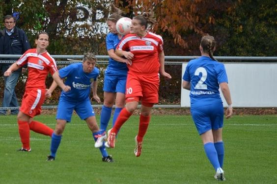 Zwei Fu&szlig;ballspielerinnen springen um den Ball, w&auml;hrend zwei weitere Spielerinnen in blauen Trikots in der N&auml;he stehen.