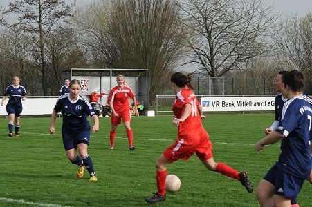 Zwei Fu&szlig;ballmannschaften spielen auf einem Rasenplatz, eine Spielerin in Rot dribbelt den Ball, w&auml;hrend andere reagieren.