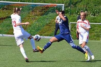 Zwei Fu&szlig;ballspielerinnen im Wettkampf um den Ball auf einem Kunstrasenplatz, eine Spielerin in Blau, die andere in Wei&szlig;.