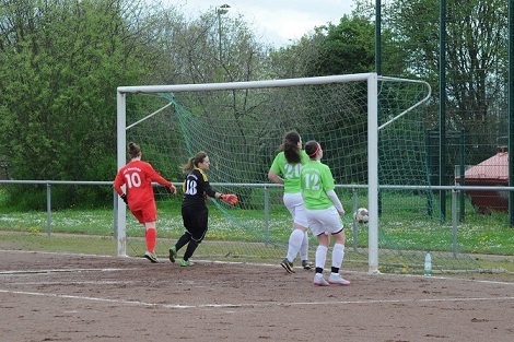 Zwei Fu&szlig;ballspielerinnen in gr&uuml;nen Trikots reagieren auf einen Schuss, w&auml;hrend die Torfrau im roten Dress bereitsteht.
