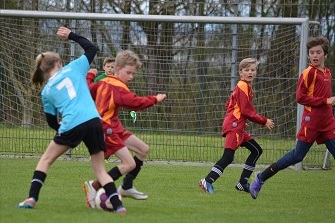 Vier Kinder spielen Fu&szlig;ball auf einem Rasenplatz, der Torwart in Blau dreht sich zum Ball.