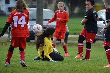 M&auml;dchenfu&szlig;ballspiel: Torh&uuml;terin in gelber Kleidung sitzt auf dem Rasen, w&auml;hrend Spielerinnen um den Ball k&auml;mpfen.