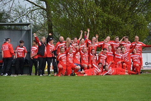 Feiernde Frauenfu&szlig;ballmannschaft in roten Trikots posiert auf dem Rasen, einige Heben die Arme freudig hoch.