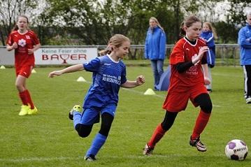 Zwei M&auml;dchen in Sportkleidung laufen w&auml;hrend eines Fu&szlig;ballspiels auf einem gr&uuml;nen Rasenfeld hinter einem Ball.