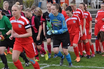 Fu&szlig;ballmannschaften in roten und schwarzen Trikots betreten das Spielfeld, eine Torh&uuml;terin h&auml;lt einen Ball in der Hand.
