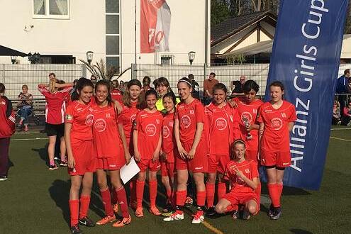 Gruppe junger Fußballerinnen in roten Trikots posiert auf einem Sportplatz mit Allianz Girls Cup-Banner im Hintergrund.