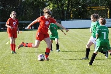 Mädchenfußballspiel auf einem Rasenplatz; Spielerin in rotem Trikot schießt den Ball, umgeben von Gegnerinnen in grünen Trikots.