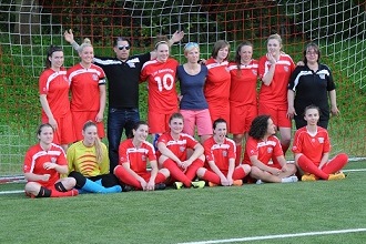 Gruppenfoto einer Fu&szlig;ballmannschaft in roten Trikots mit Trainerin auf einem Kunstrasenplatz vor dem Tor.