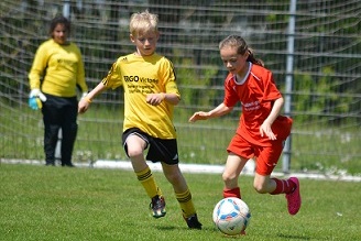 Zwei Kinder spielen Fu&szlig;ball auf einem Platz, ein Junge in gelbem Trikot und ein M&auml;dchen in rotem Trikot.