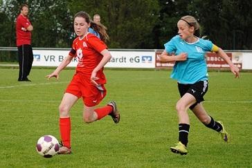 Zwei Mädchen spielen Fußball auf einem grünen Feld, eines in rotem, das andere in blauem Trikot.
