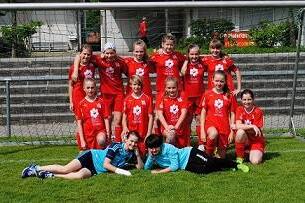 M&auml;dchenfu&szlig;ballmannschaft in roten Trikots posiert fr&ouml;hlich vor einem Fu&szlig;balltor auf gr&uuml;ner Wiese.